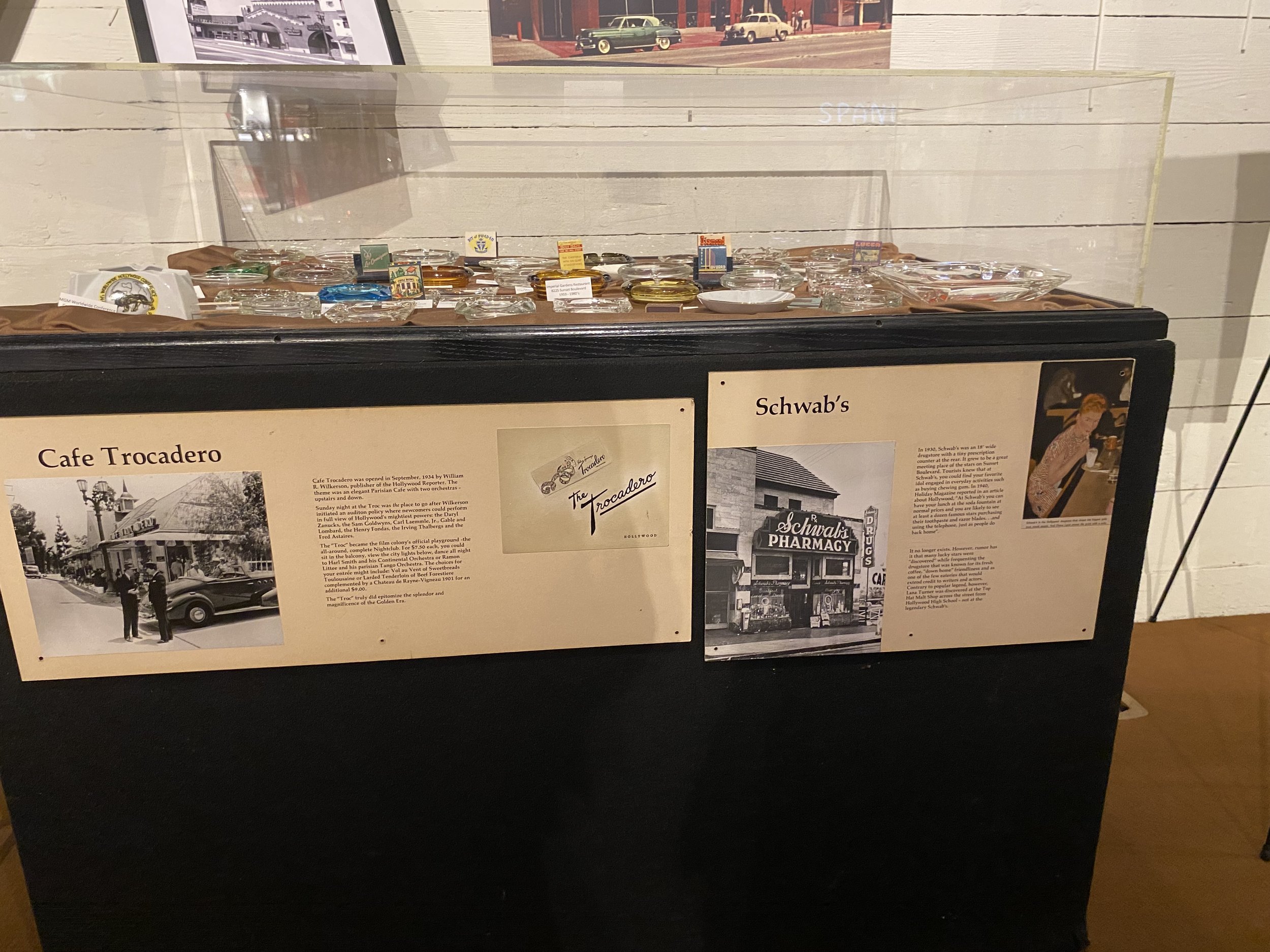 A display case containing various pins and badges, with informational posters about Cafe Trocadero and Schwab's pharmacy on a black stand.