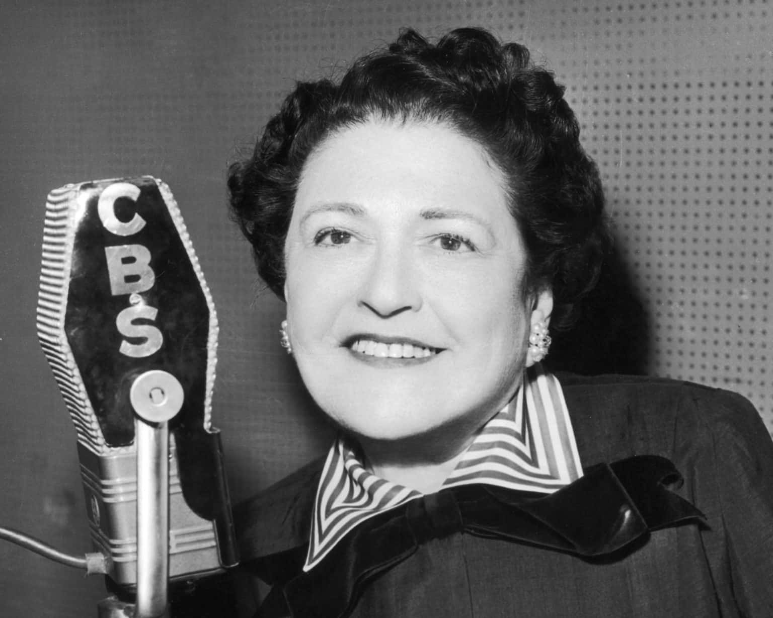 Black and white photograph of a woman with short, curly hair, smiling, wearing earrings, a striped collar, and a bow tie, standing next to a microphone labeled "CBSO".