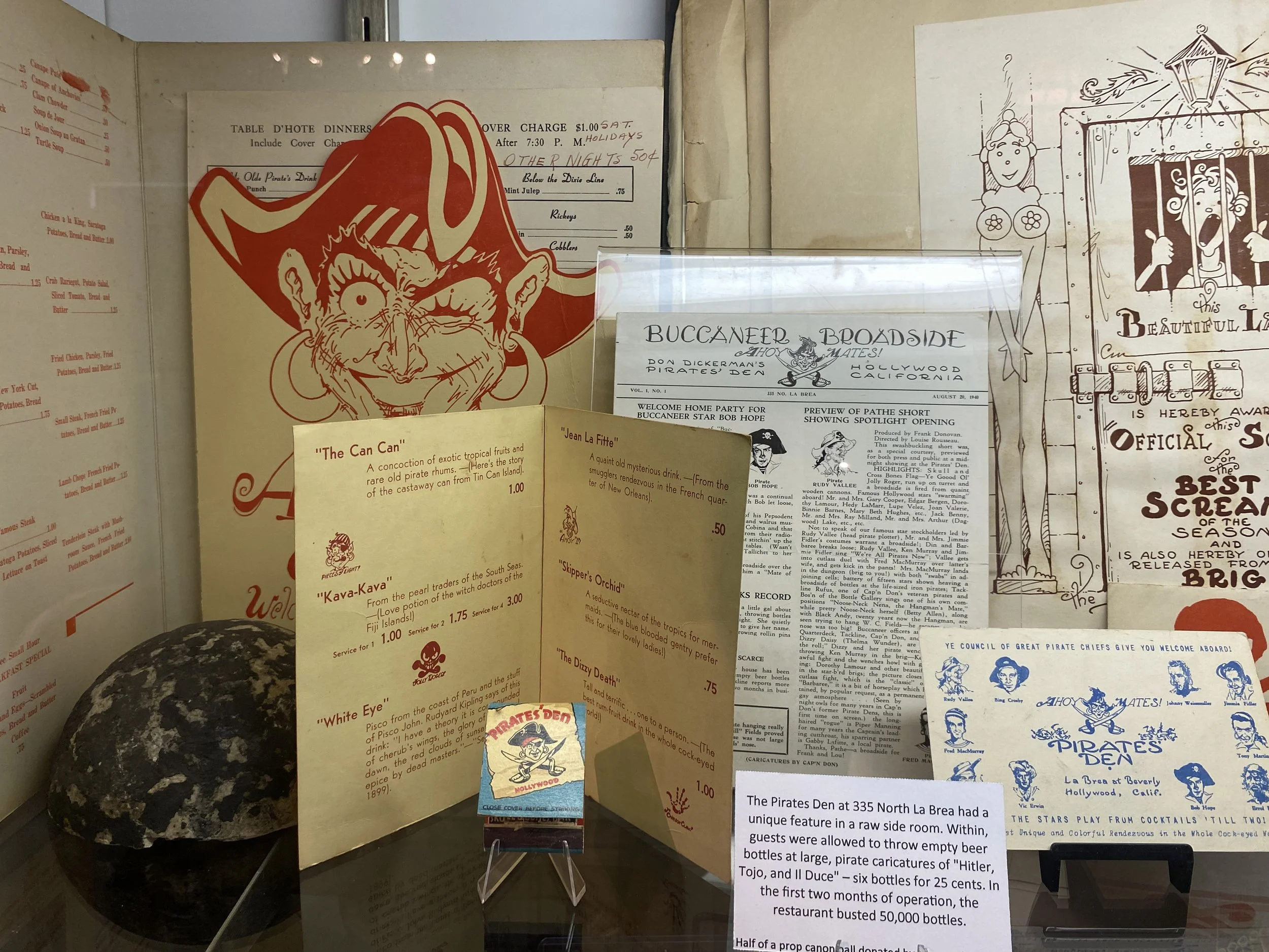 Display case with pirate-themed items including menus, caricatures, and decorations, featuring a pirate's face sticker, a rock, and informational cards about pirate stories and a pirate den in Hollywood, California.