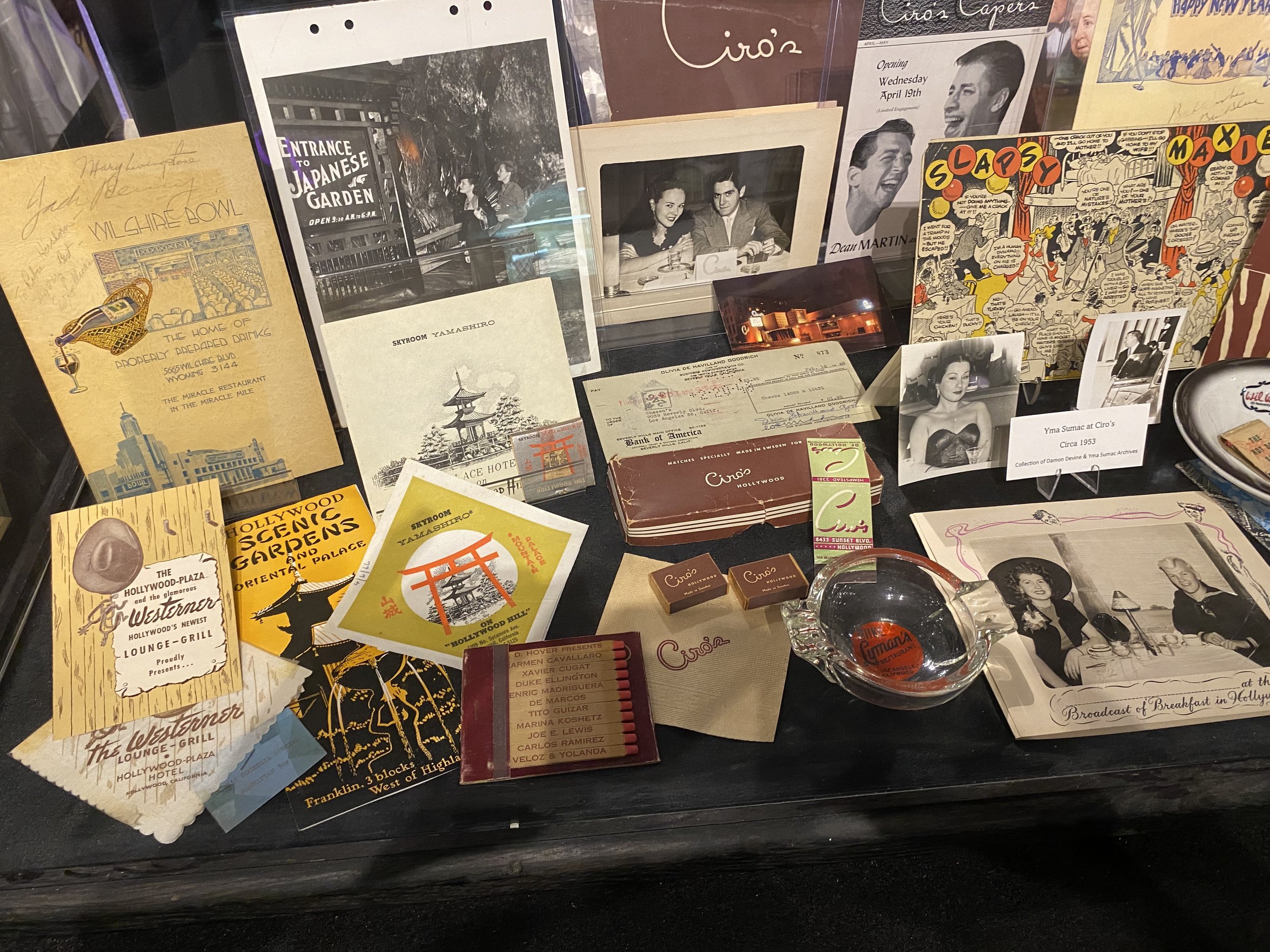 A collection of vintage photographs, postcards, and memorabilia on display, including black and white photos of people, vintage flyers, a souvenir ashtray, and small souvenirs related to Ciro's, a historic Hollywood nightclub.