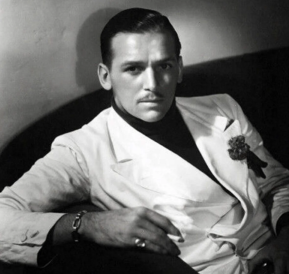 Black and white photo of a man wearing a white suit with a brooch, sitting on a dark sofa, looking at the camera.