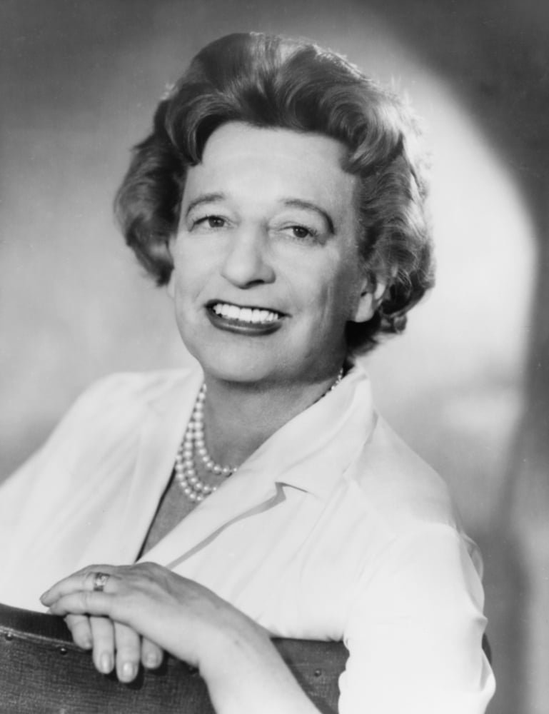Black and white portrait of an elderly woman with curled hair, smiling, wearing a pearl necklace, a white blouse, and a ring on her finger.