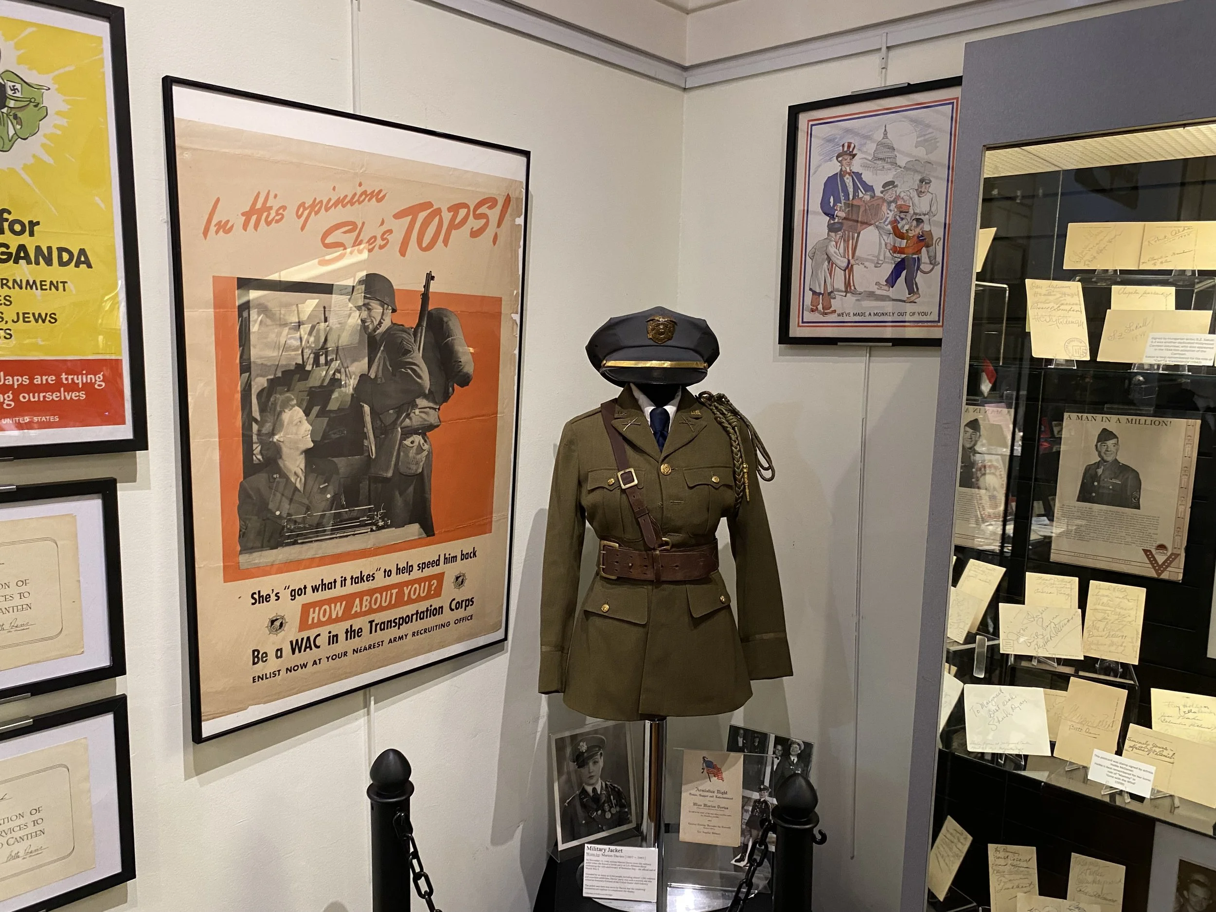 Display in a museum featuring a World War II military uniform on a mannequin, vintage posters, and framed documents on the walls and in a glass case.