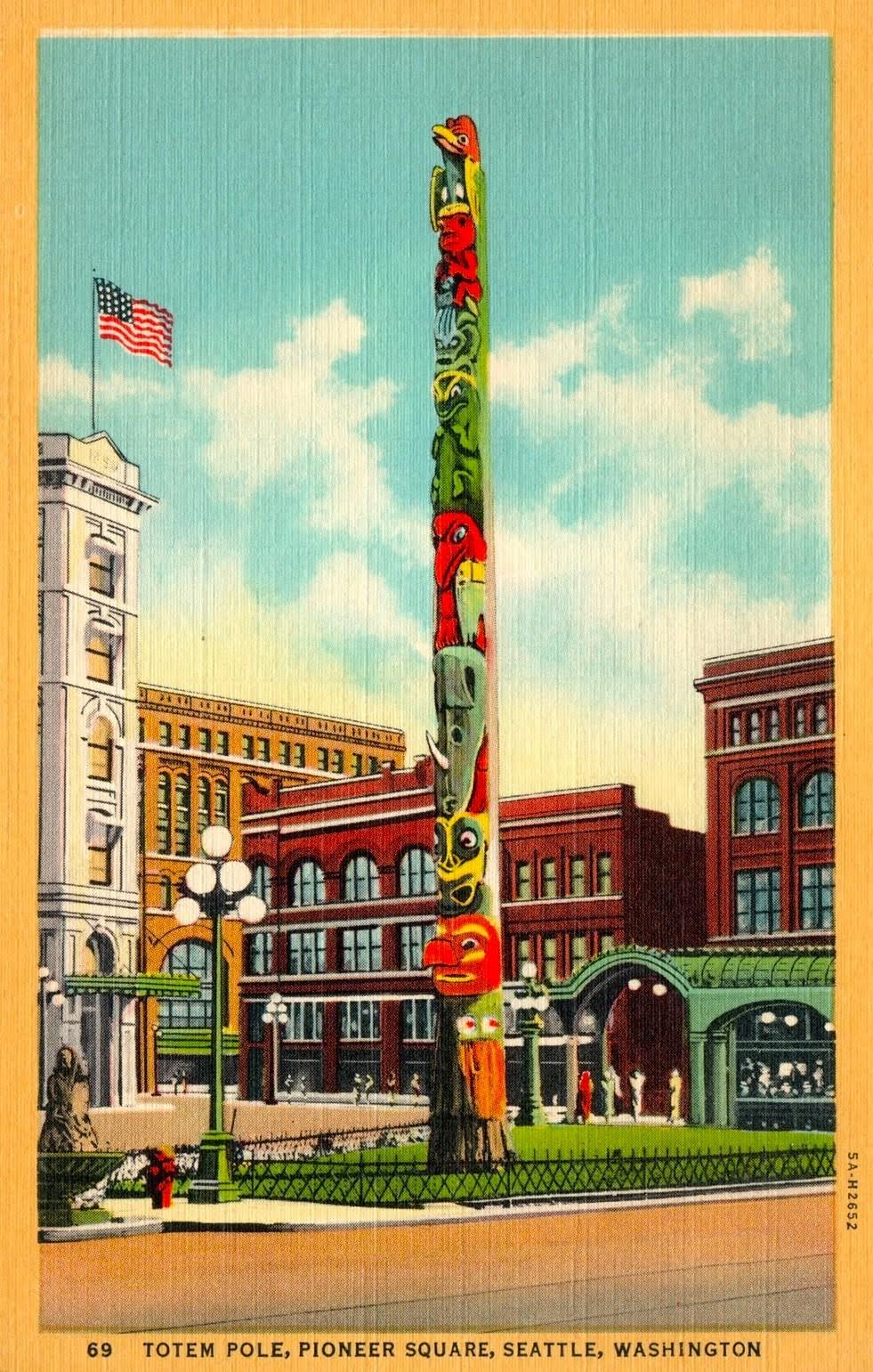 Colorful totem pole standing in Pioneer Square park, Seattle, Washington, with historic buildings, a flag, and a clear sky in the background.