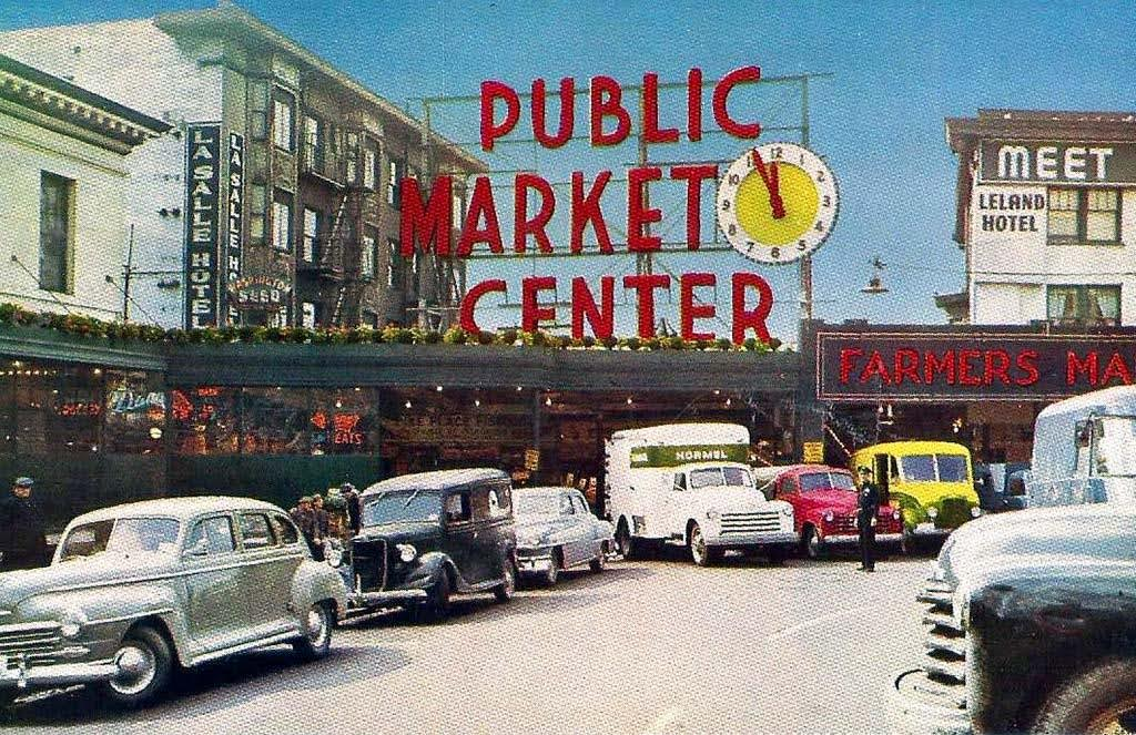 Vintage photograph of a public market center with cars parked in front, a large red sign above reading 'Public Market Center' with a clock, and other shop signs such as 'Farmers Market' and 'Leland Hotel' visible on surrounding buildings.