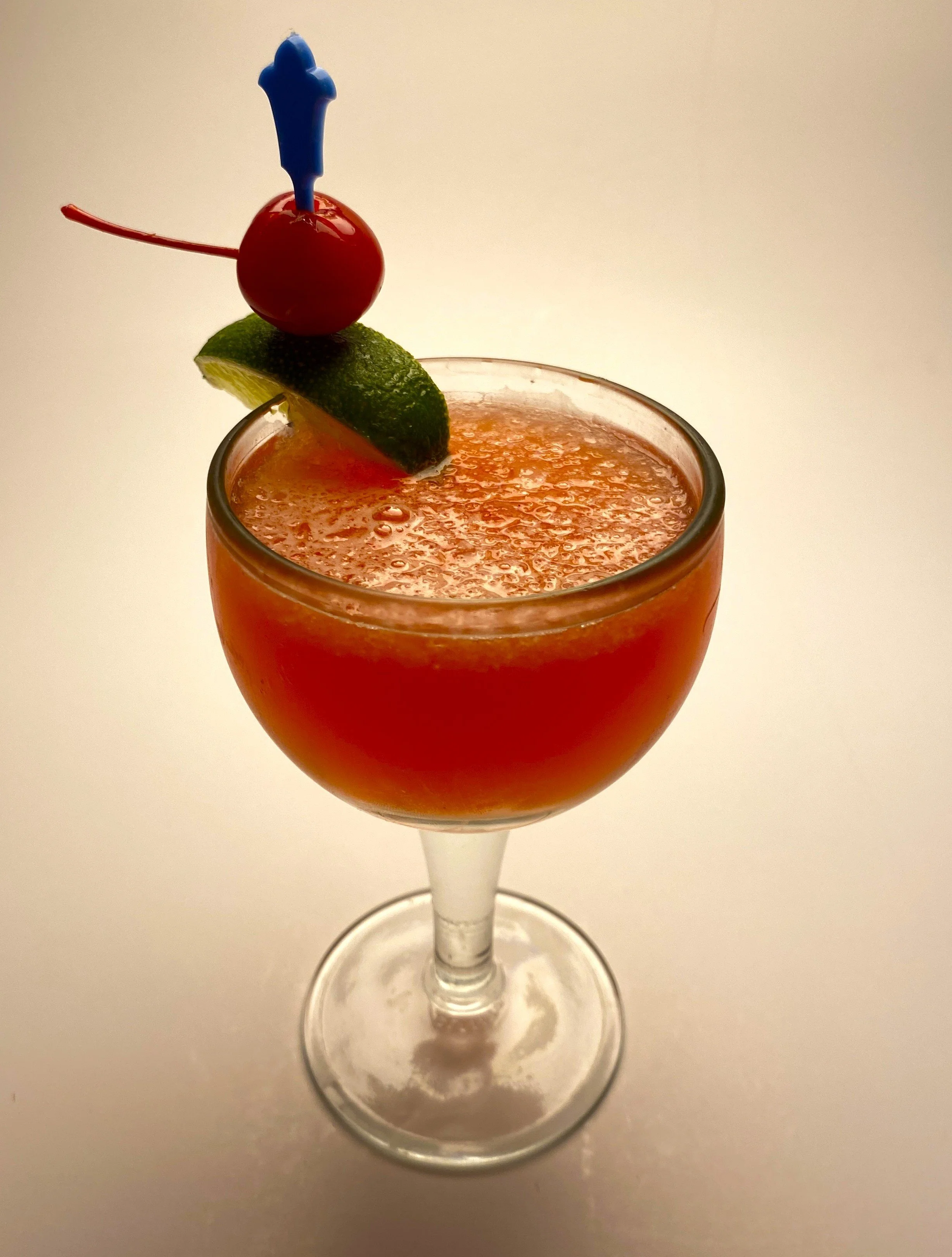 A cocktail drink in a stemmed glass garnished with a cherry, lime wedge, and blue plastic pick.