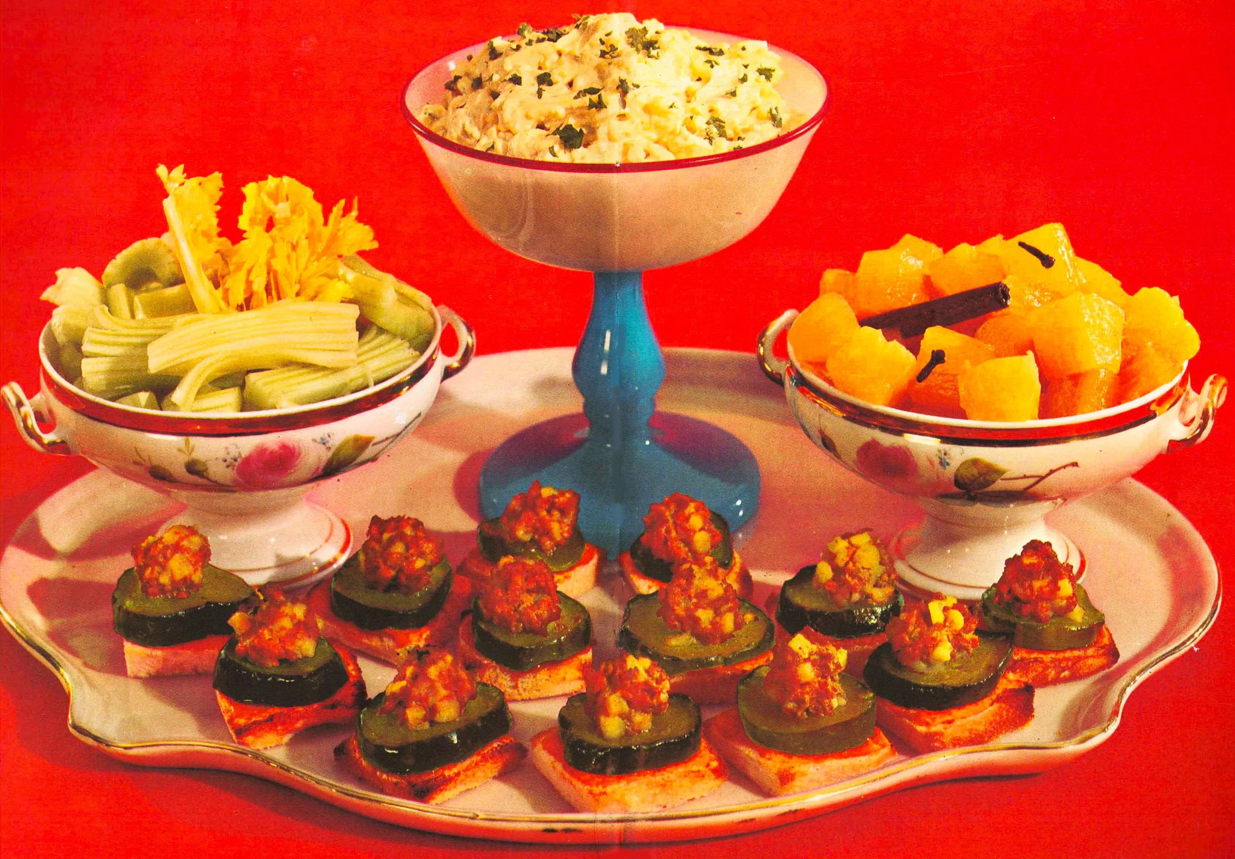Assorted appetizers displayed on a decorative tray, including cucumber and veggie bites with toppings, and bowls of pasta salad, cubed melon, and cubed butternut squash, on a red background.
