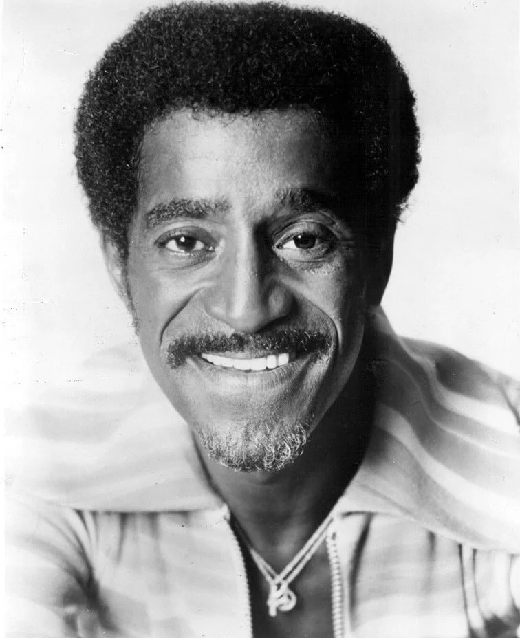 Black and white portrait of a man with a mustache and curly hair, smiling, wearing a striped shirt and a necklace.
