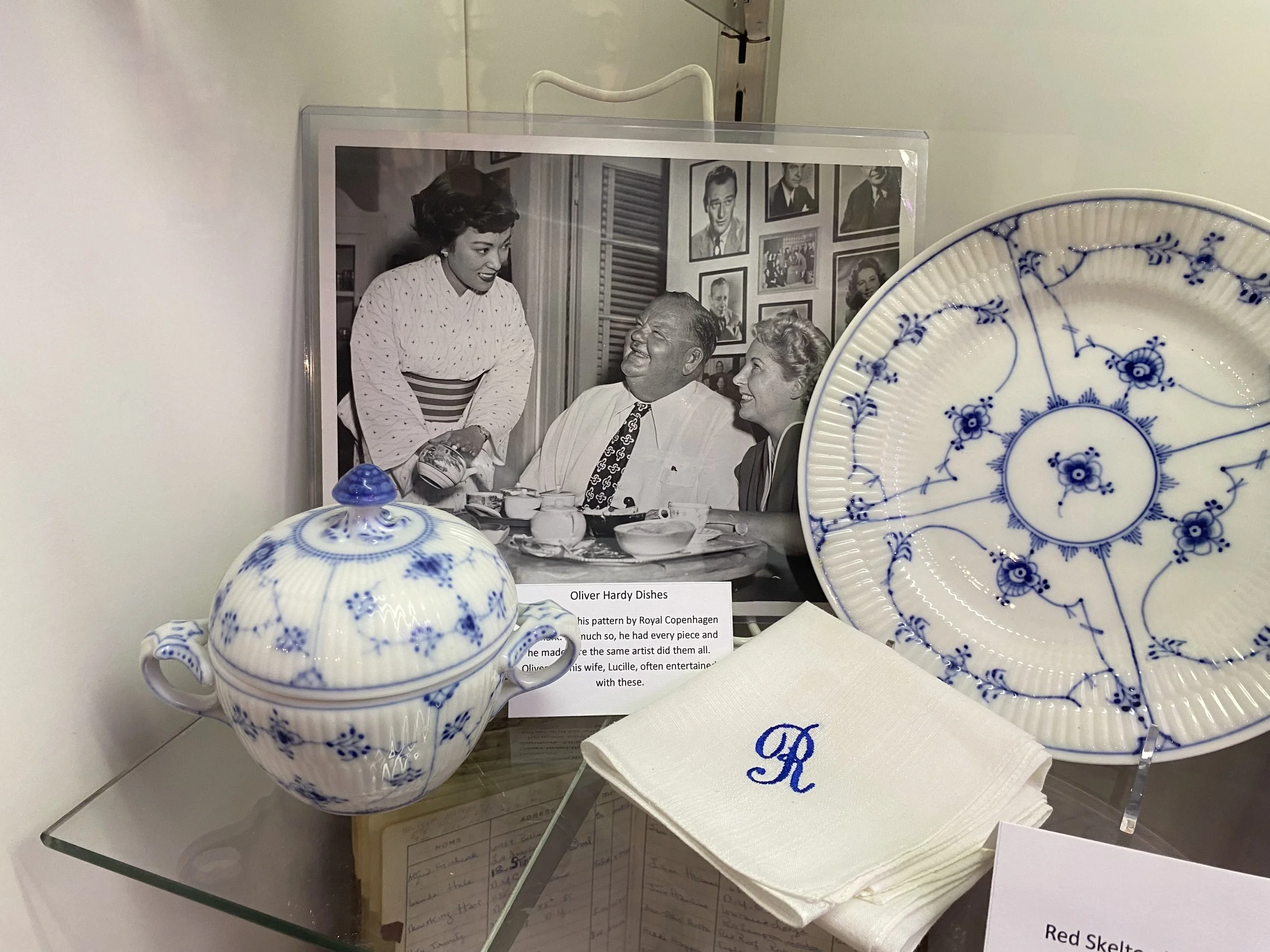 A collection of blue and white porcelain dishes including a sugar bowl and a plate, a set of white cloth napkins embroidered with a blue letter R, accompanied by a black-and-white photograph of three people dining and engaging in conversation, displa