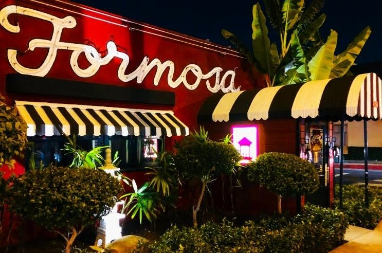 Nighttime view of a restaurant or cafe named 'Formosa' with a large neon sign, striped awnings, and surrounded by green plants and trees.