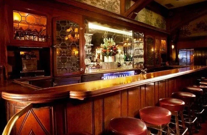 Empty wooden bar with bar stools in a dimly lit, vintage-style pub or restaurant, featuring a back bar with glassware and a floral arrangement.
