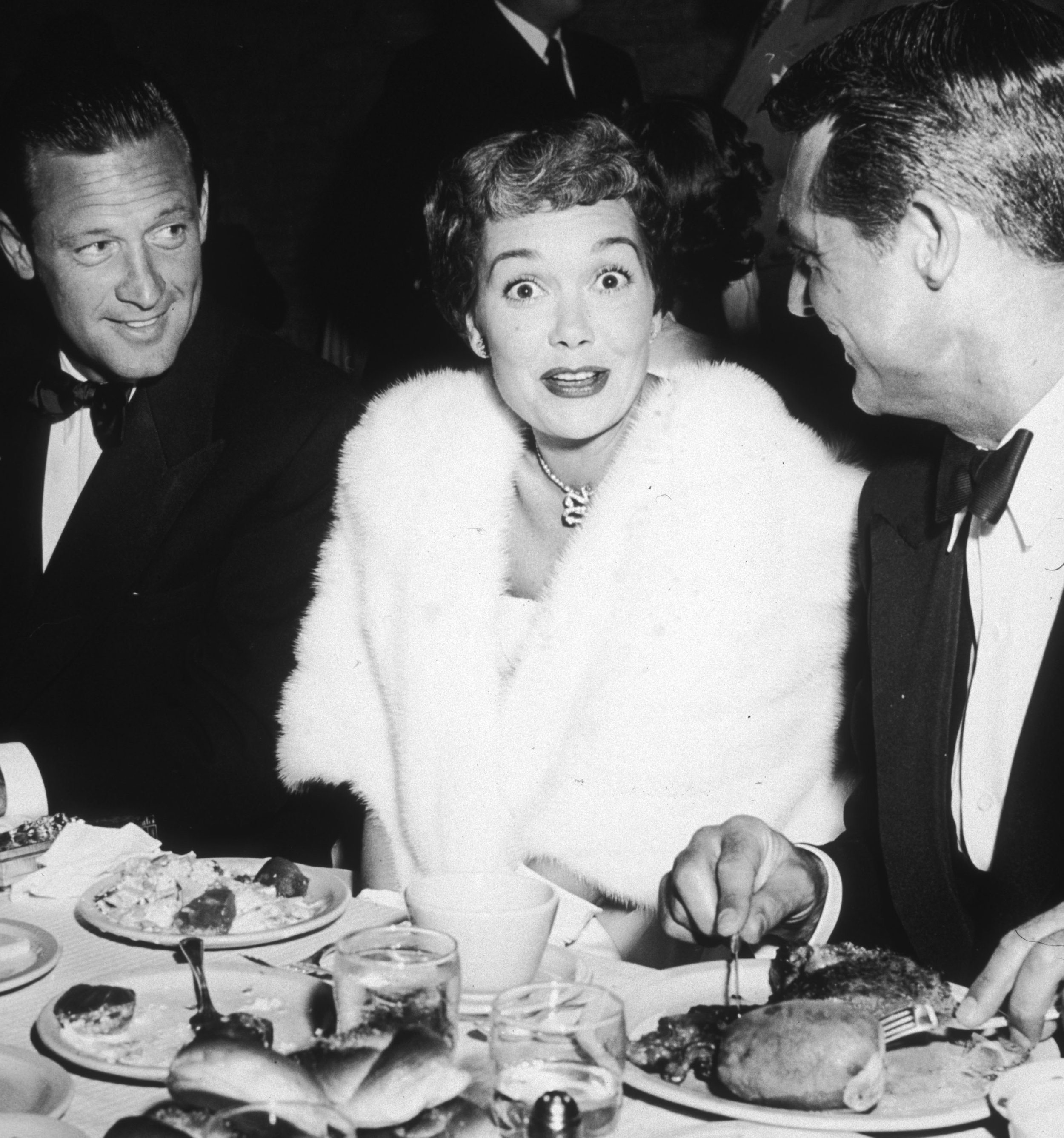 A woman in a white fur stole sitting at a dinner table with two men in tuxedos, appearing surprised and engaged in conversation, with food and drinks on the table.