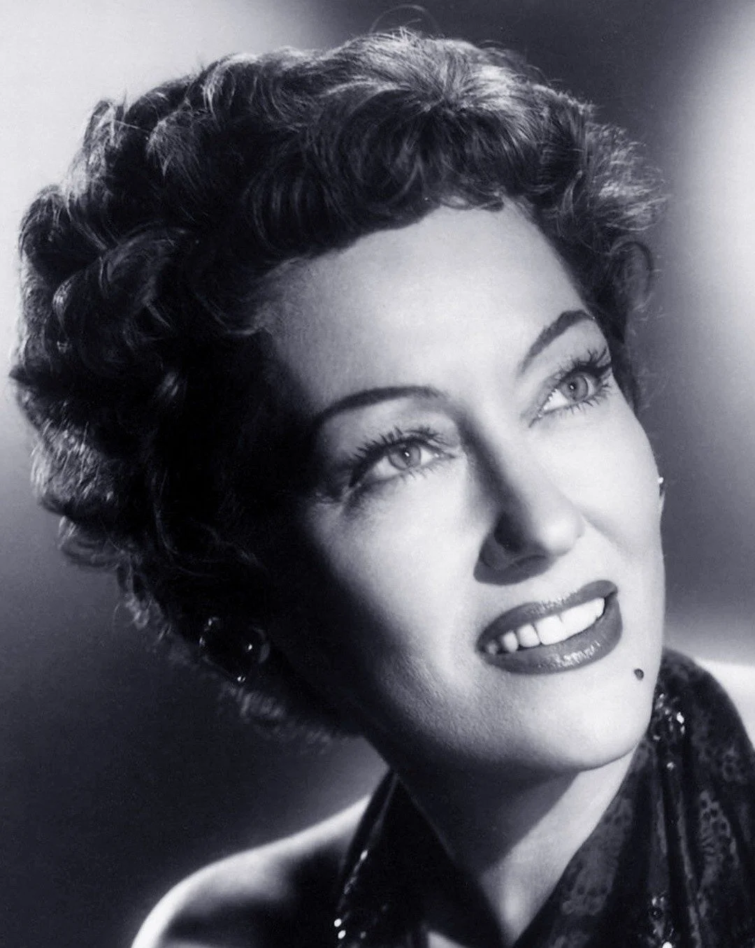 Black and white portrait of a woman with short, curly hair, bright eyes, and a beauty mark above her lips, smiling slightly and looking upwards.