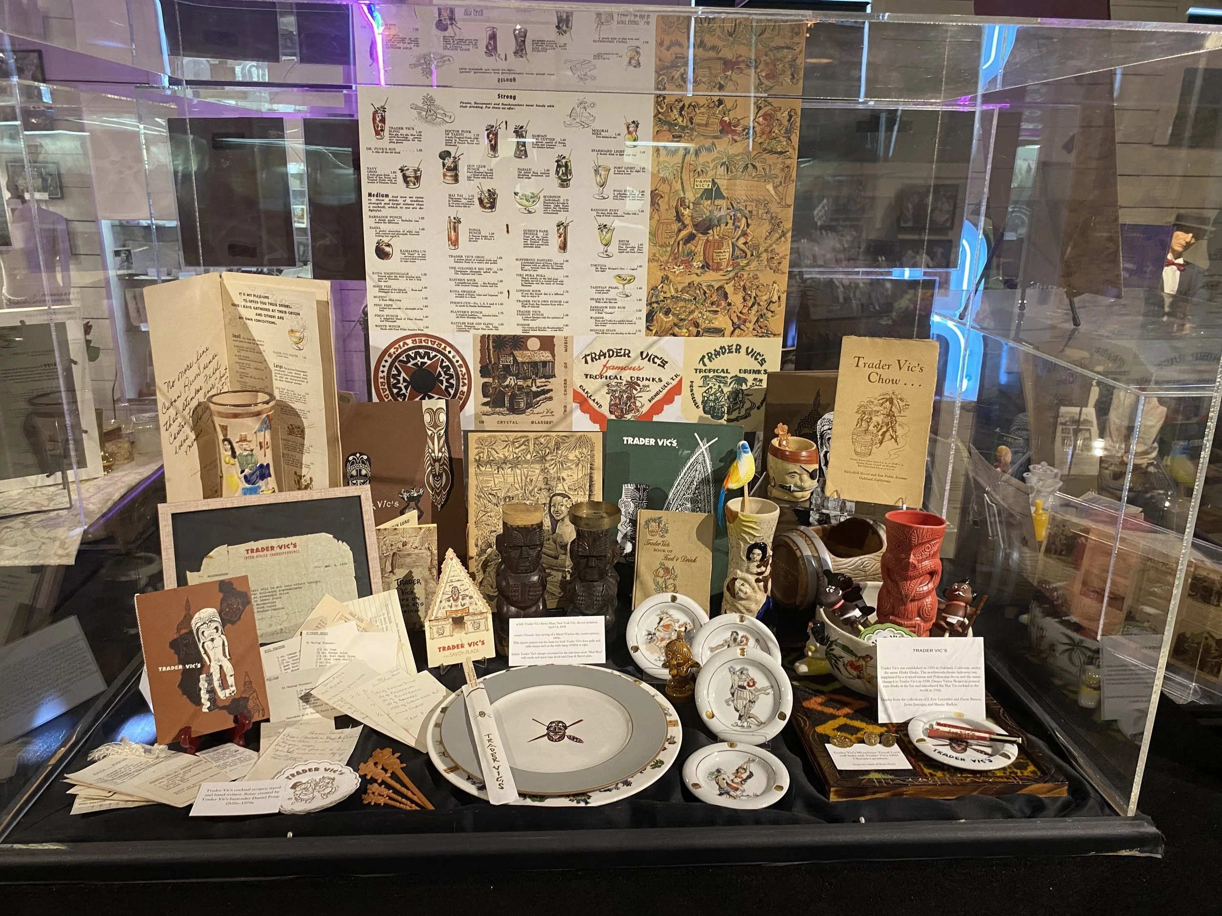 A display case with various James Michener collectibles, such as pottery, plates, artwork, booklets, and memorabilia related to Trader Vic's and Polynesian themes.