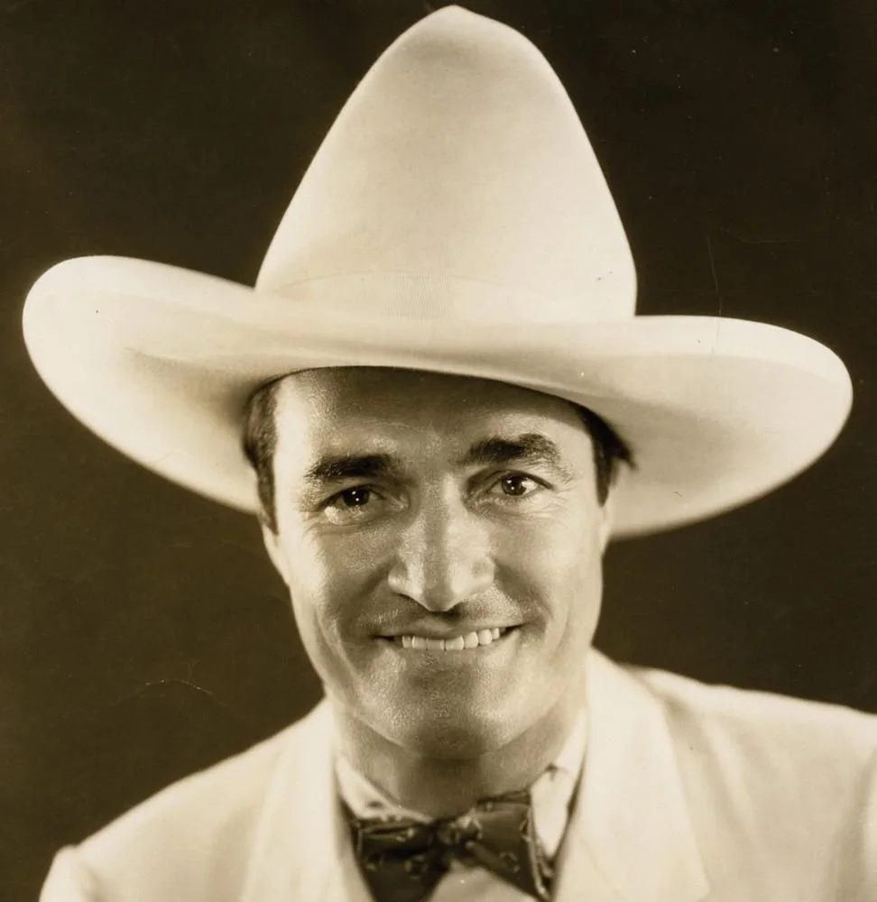 Black and white portrait of a man wearing a large cowboy hat, a light-colored suit, and a patterned bow tie, smiling at the camera.