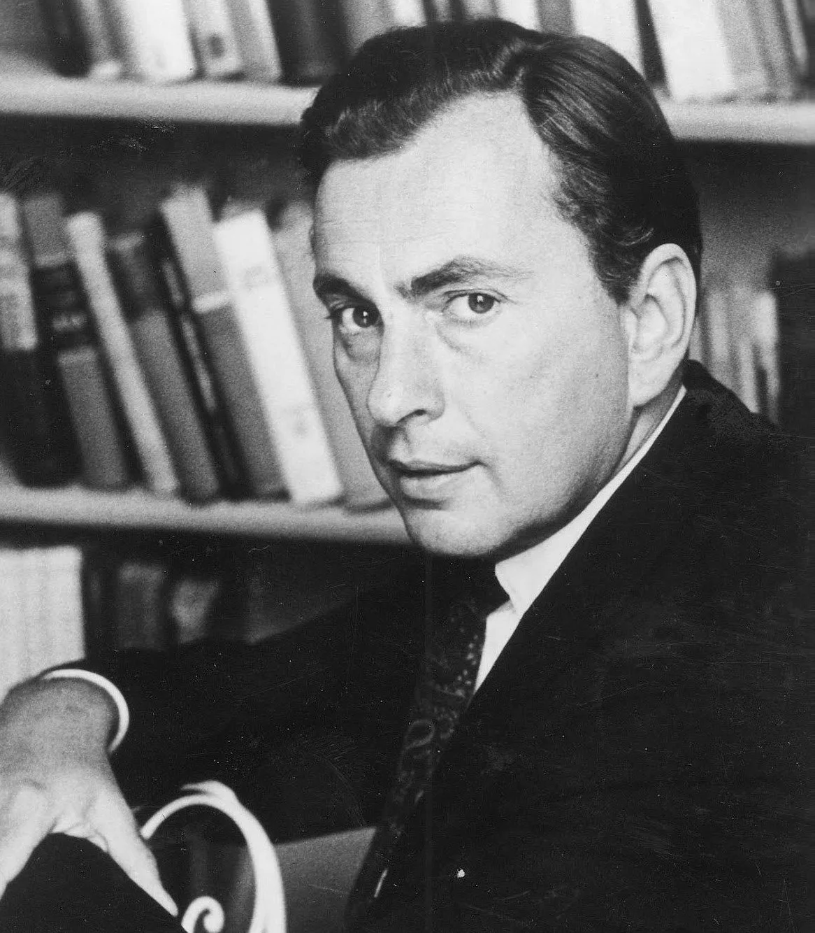 Black and white photo of a man with dark hair in a suit, sitting at a desk surrounded by books, looking directly at the camera.