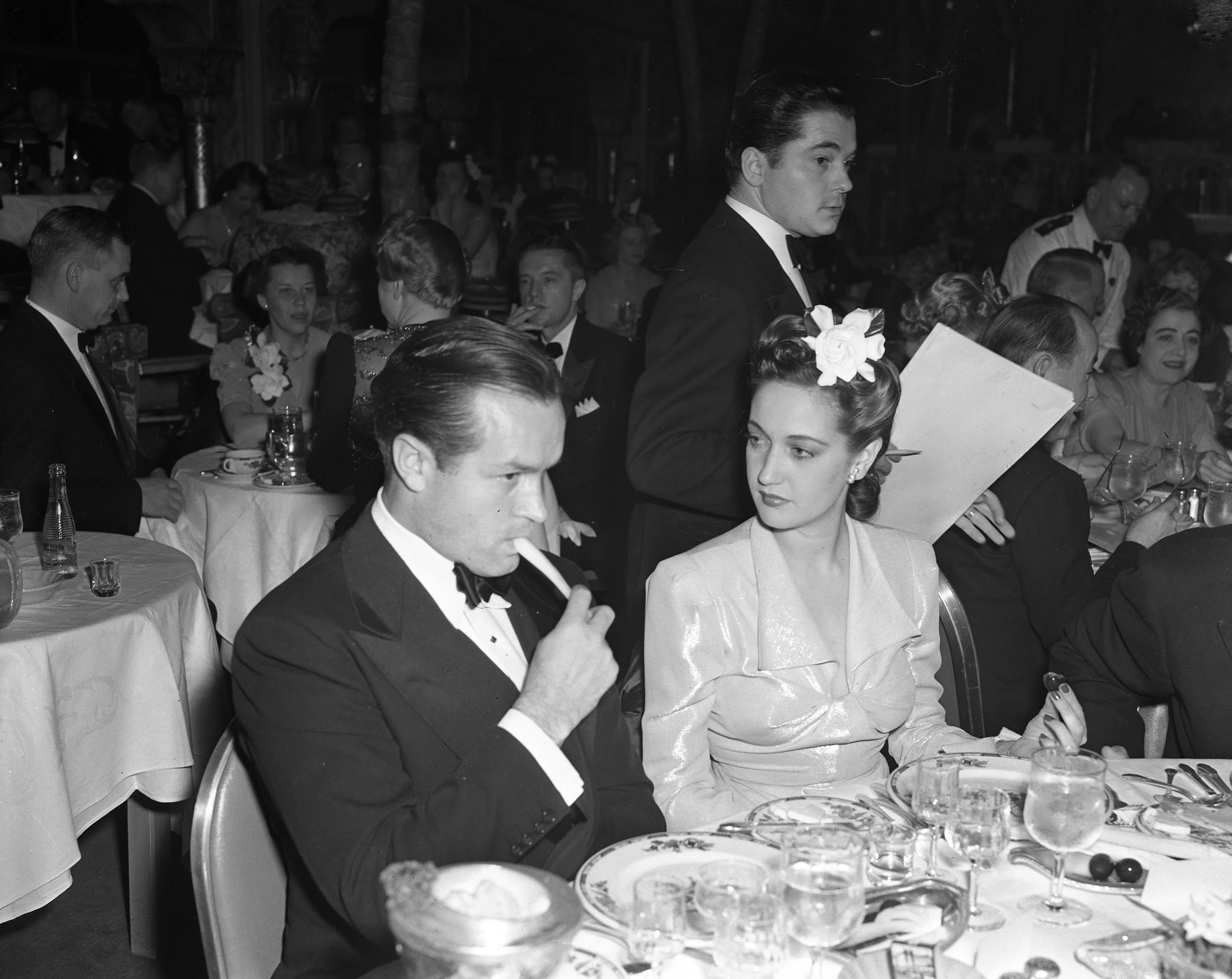 A black-and-white photo of men and women in formal attire at a dining event. One man is smoking a cigarette and sitting at a table with a woman. Others are sitting or standing in the background, engaged in conversation.