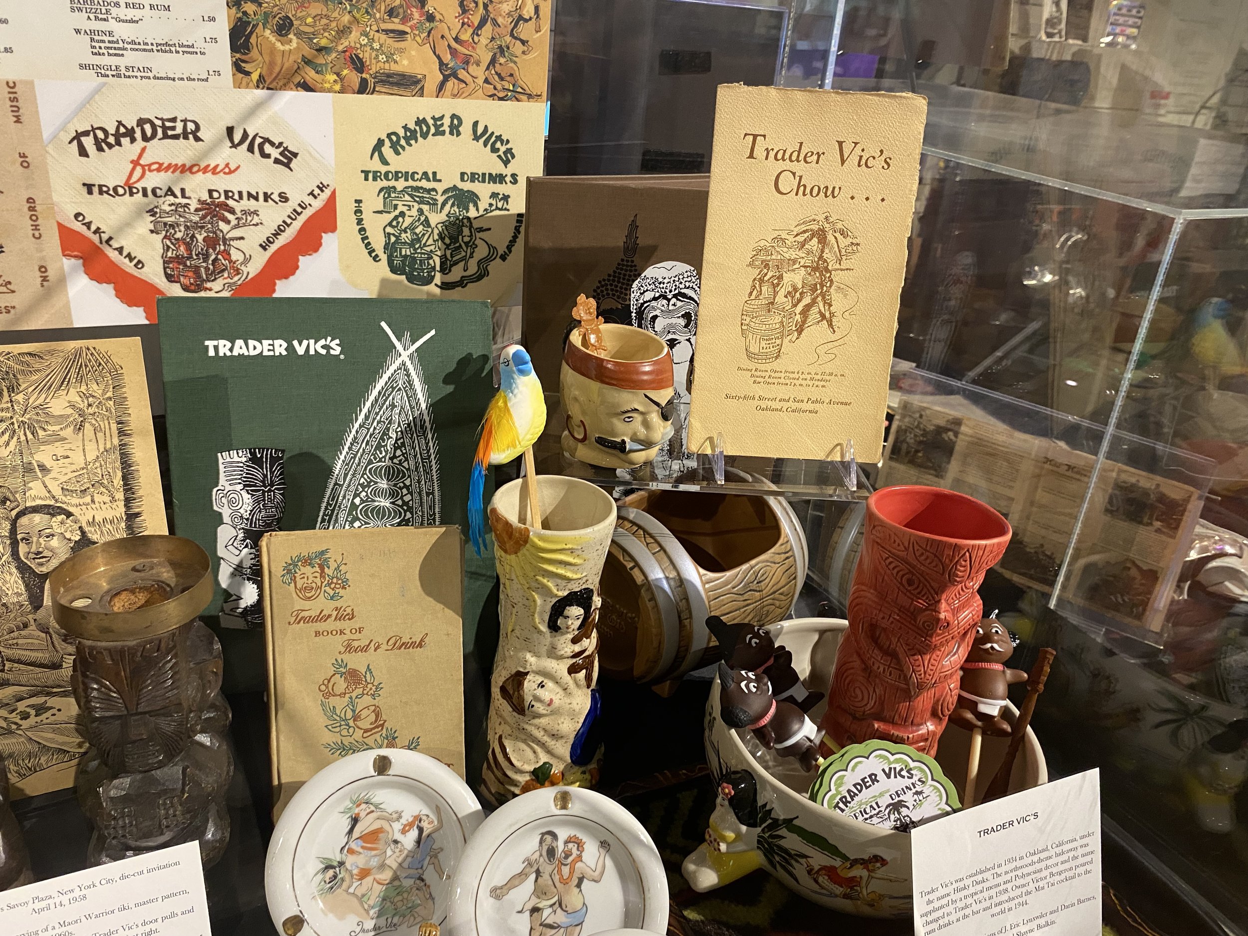 Display of Trader Vic's souvenirs, including books, mugs, figurines, and decorative items, with Hawaiian and tiki themes.