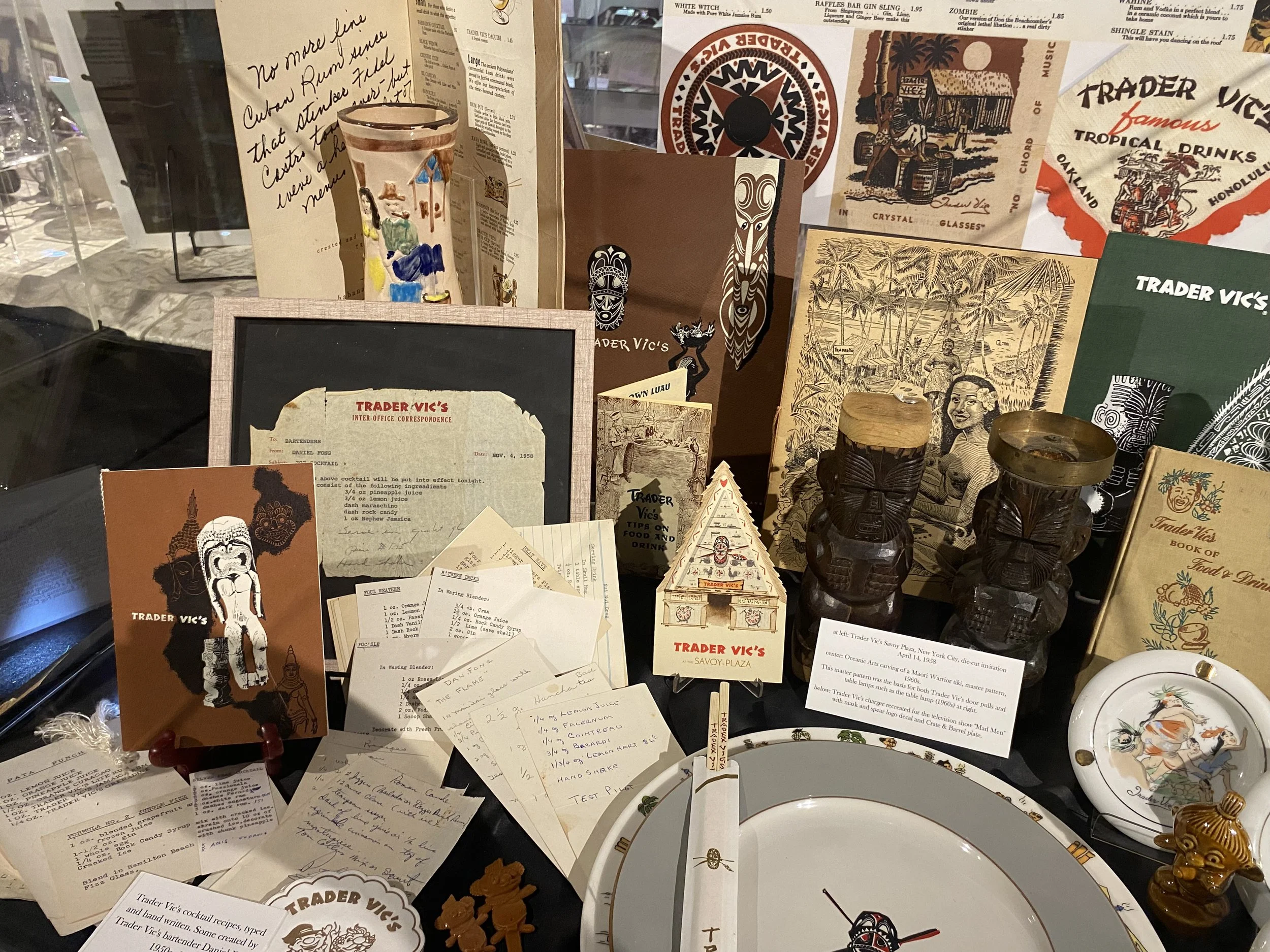 A display case featuring vintage Trader Vic's memorabilia, including old menus, photographs, tiki mugs, carved wooden figurines, and decorative plates.