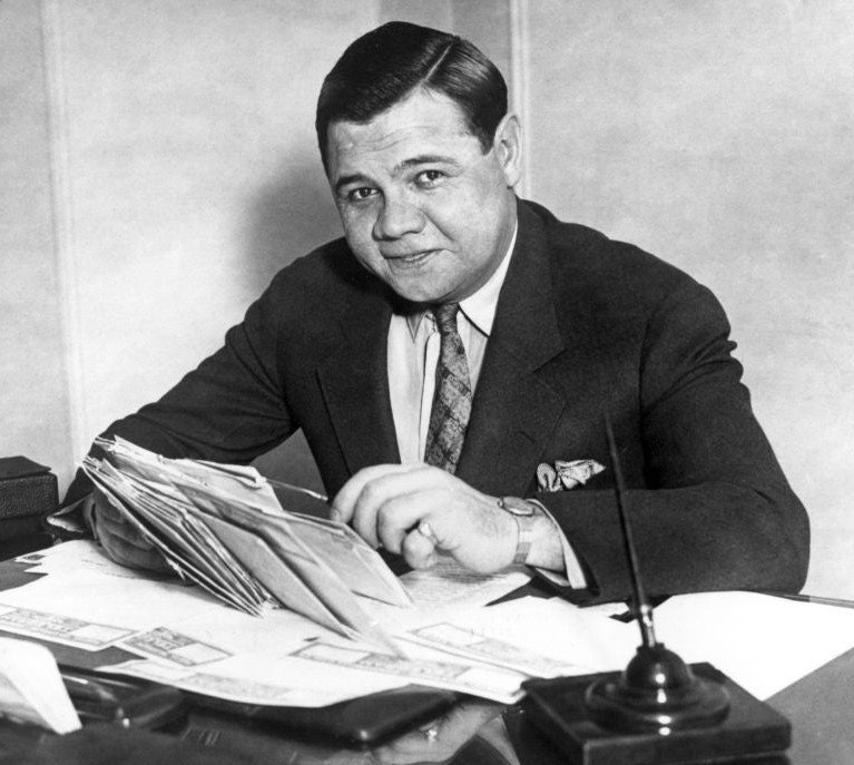 Black and white photo of a man in a suit sitting at a cluttered desk with papers and files, smiling at the camera.