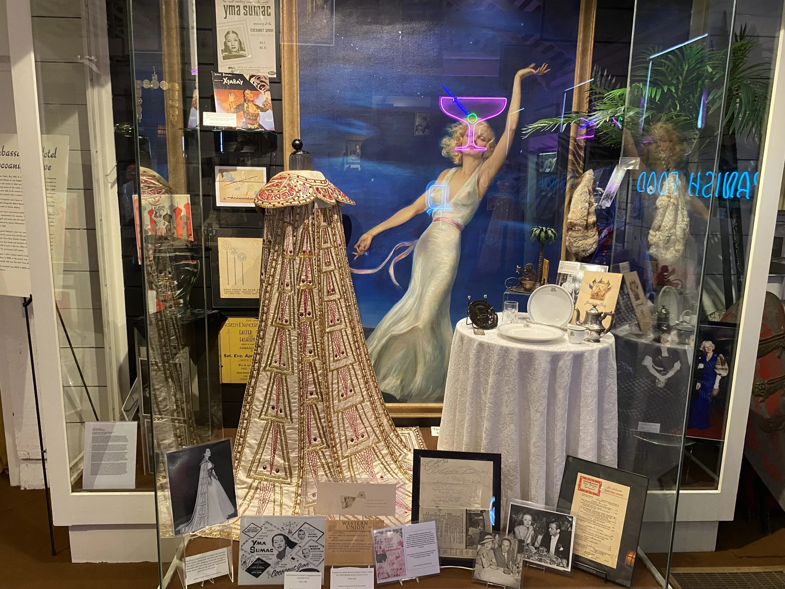 A museum display case featuring vintage items, including a large beaded dress, a white table with dishes and silverware, a painting of a woman in a white dress with a cocktail glass on her face, various photographs, documents, and artifacts related t