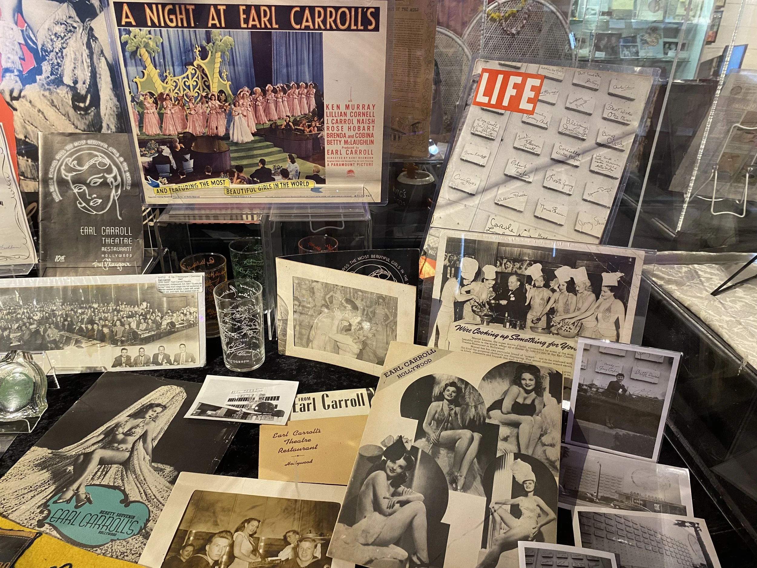 Display of vintage photographs, posters, and memorabilia related to Earl Carroll, including a sign for 'A Night at Earl Carroll's,' black-and-white pictures of women in swimwear and costumes, a 'LIFE' magazine display, signed photographs, and various