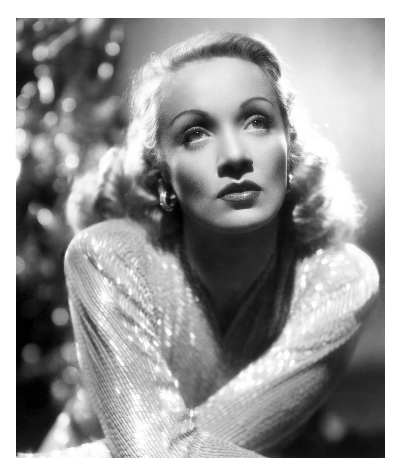 Black and white portrait of a woman with styled hair and makeup, looking upward with a pensive expression, wearing earrings and a shimmering outfit.