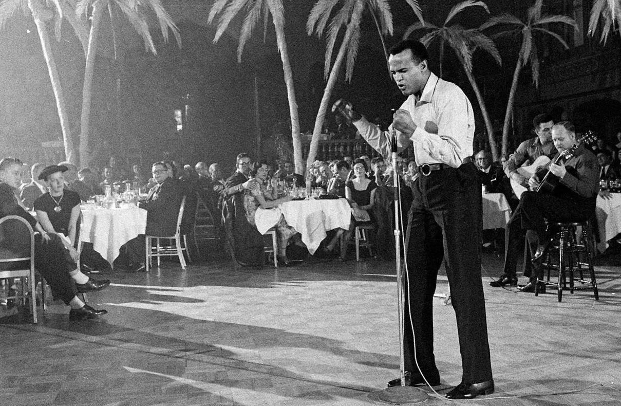 A black and white photo of a singer and guitarist performing at a restaurant or club with an audience watching, surrounded by trees.