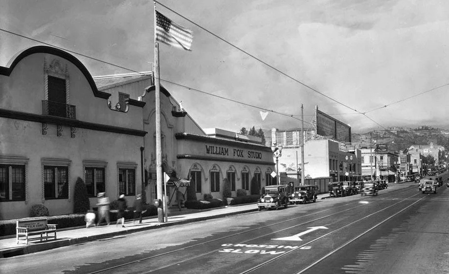 A black and white photo of a street scene in what appears to be an early to mid-20th century town. There are vintage cars parked along the curb and a few pedestrians walking on the sidewalk. A flag is flying on a pole, and there is a building labeled "William Fox Studio" with a marquee over the entrance. The street has tracks for streetcars, and there are some hills visible in the background.