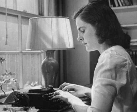A woman typing on a vintage typewriter in a room with a bookshelf and a table lamp.