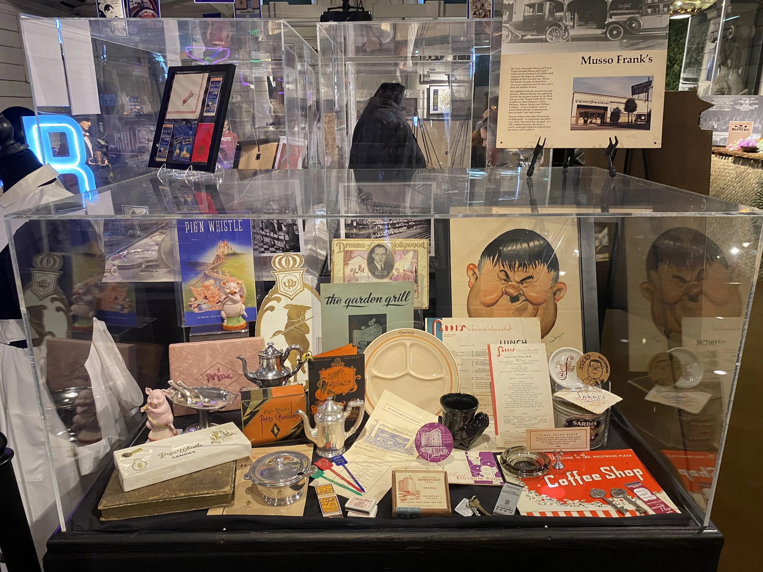 A display case containing various vintage and collectible items including small figurines, dishes, signs, posters, and a coffee shop menu. The background shows a museum or exhibit space with informational panels.
