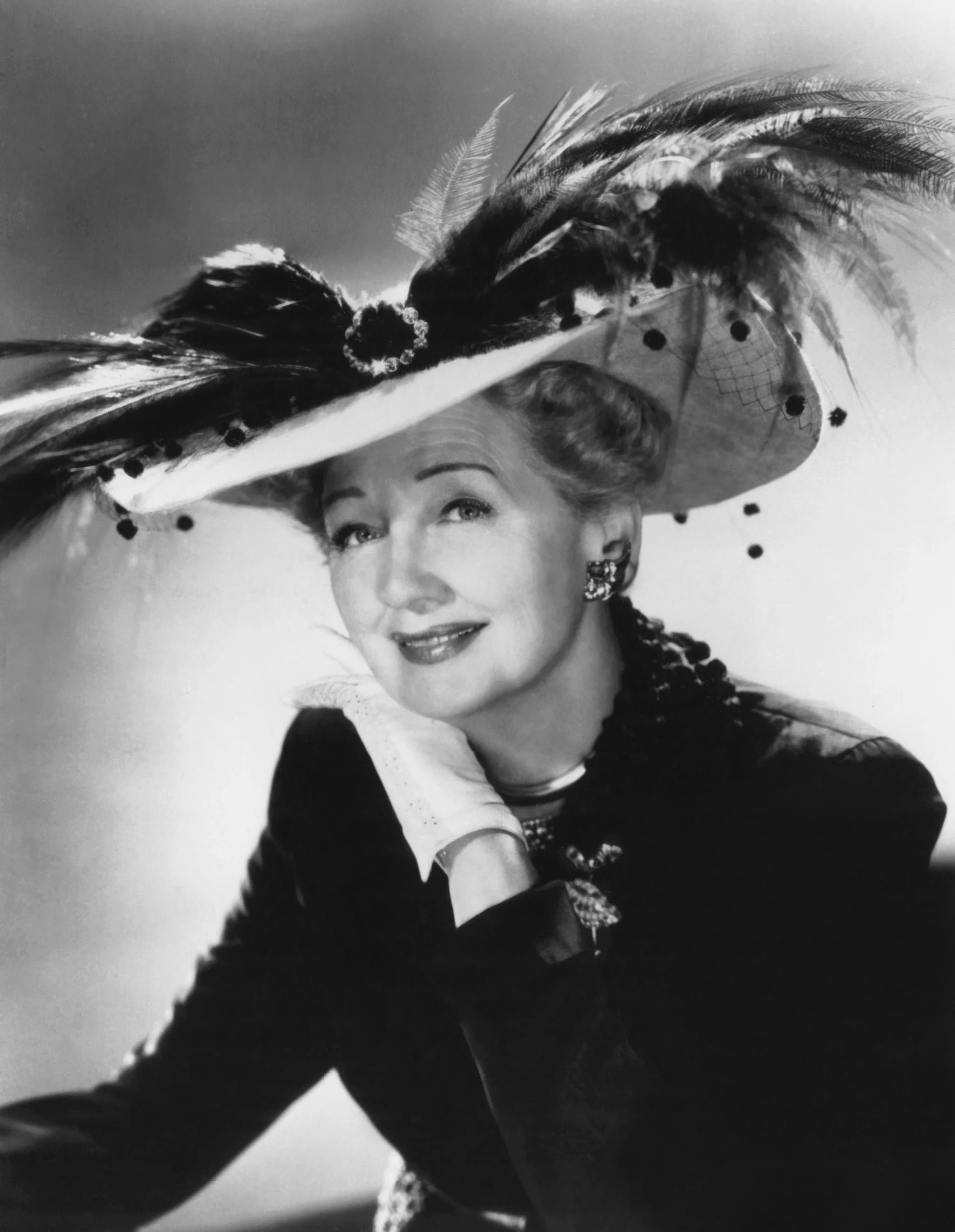 Black and white portrait of a woman wearing a large, elaborate feathered hat, with her hand resting on her chin, smiling subtly at the camera.