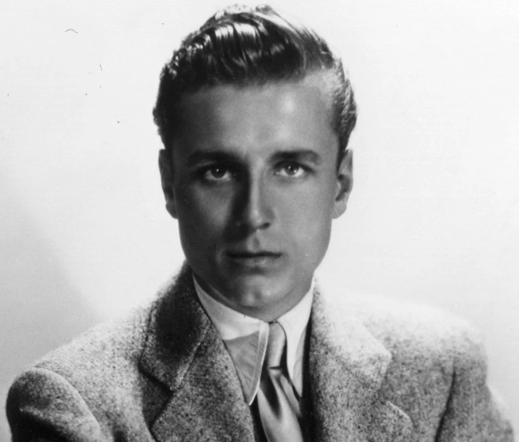 Black and white portrait of a young man with slicked-back hair wearing a suit jacket and tie.