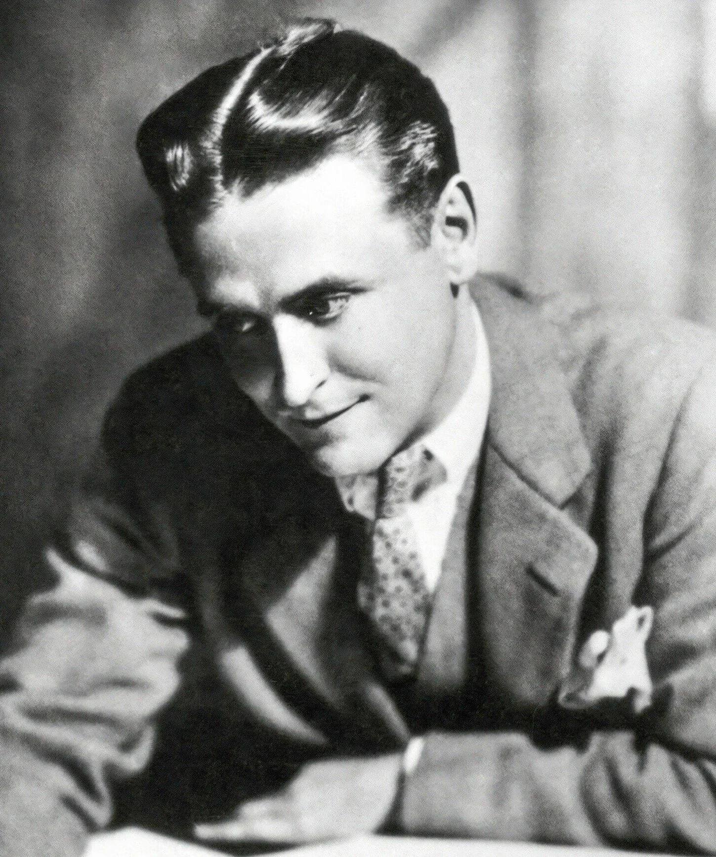 A black and white photograph of a young man with slicked-back hair, wearing a suit and tie, posing with a slight smile.