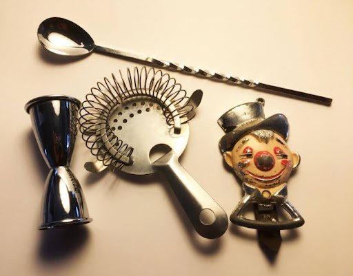 Collection of bar tools and a clown figurine, including a cocktail spoon, a jigger, a cocktail strainer, and a clown ornament with a bow tie.