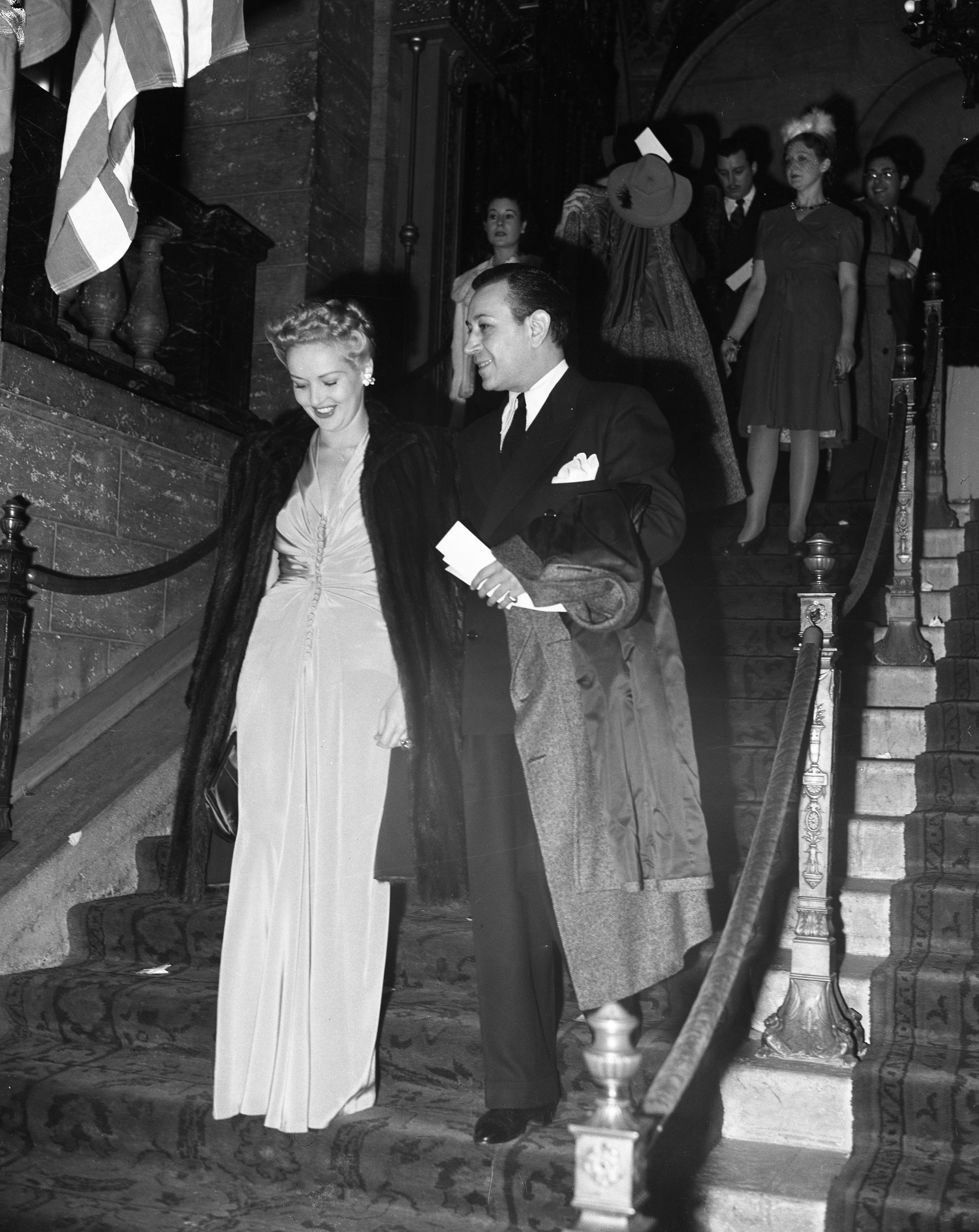 A black and white photo of a man and woman descending a staircase at a formal event, with other guests on the stairs behind them. The woman wears a long gown and fur coat, and the man is in a suit and overcoat.