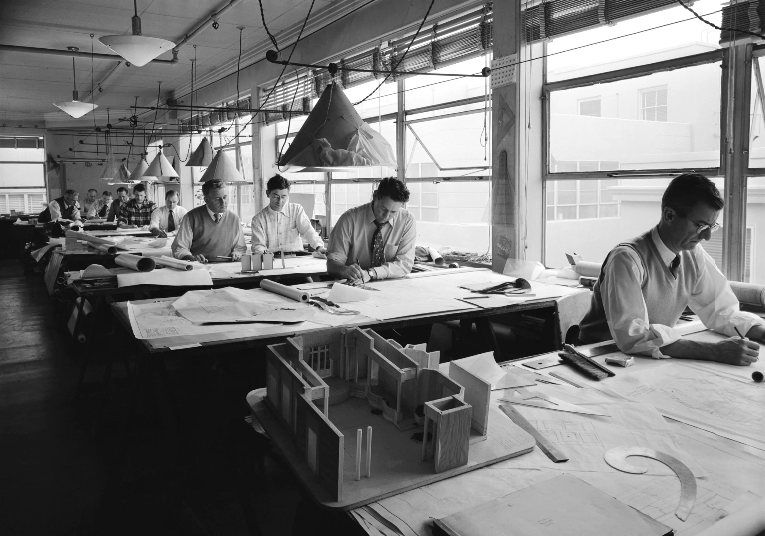 Black and white photo of an early 20th-century architecture studio with multiple men working at drafting tables, blueprints, and architectural models, large windows letting in natural light.