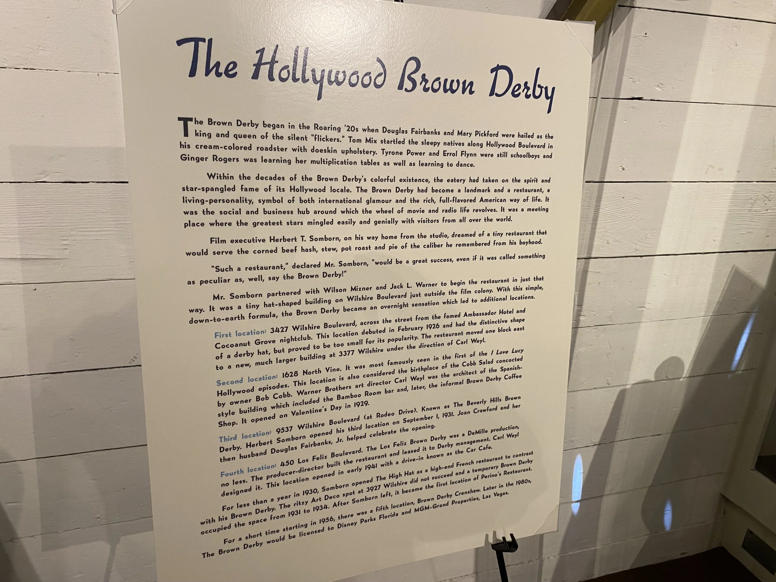 A white sign with purple and black text titled "The Hollywood Brown Derby" explaining the history of the restaurant, including its origins, various locations, and notable facts.