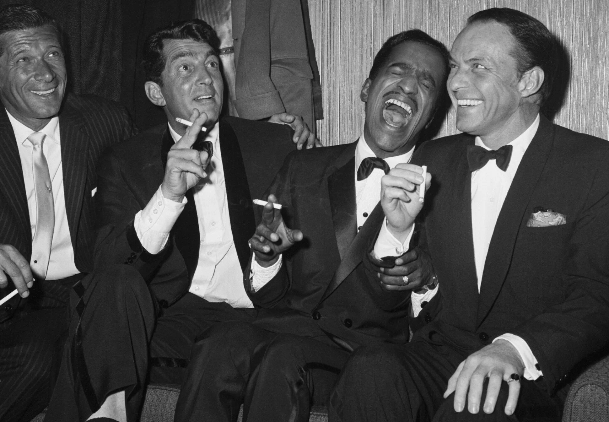 Black and white photo of five well-dressed men in tuxedos, laughing and holding cigarettes, in a social setting.