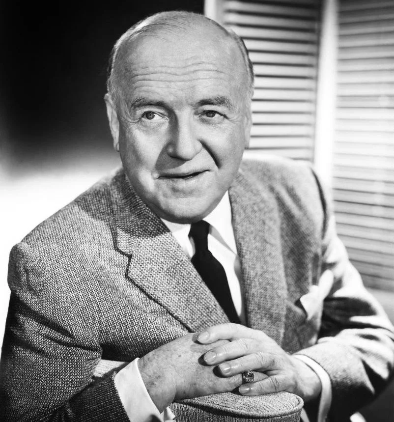 Black and white portrait of an older man with a suit and tie, resting his elbow on a surface and holding a hat in his hand, with a window with blinds in the background.