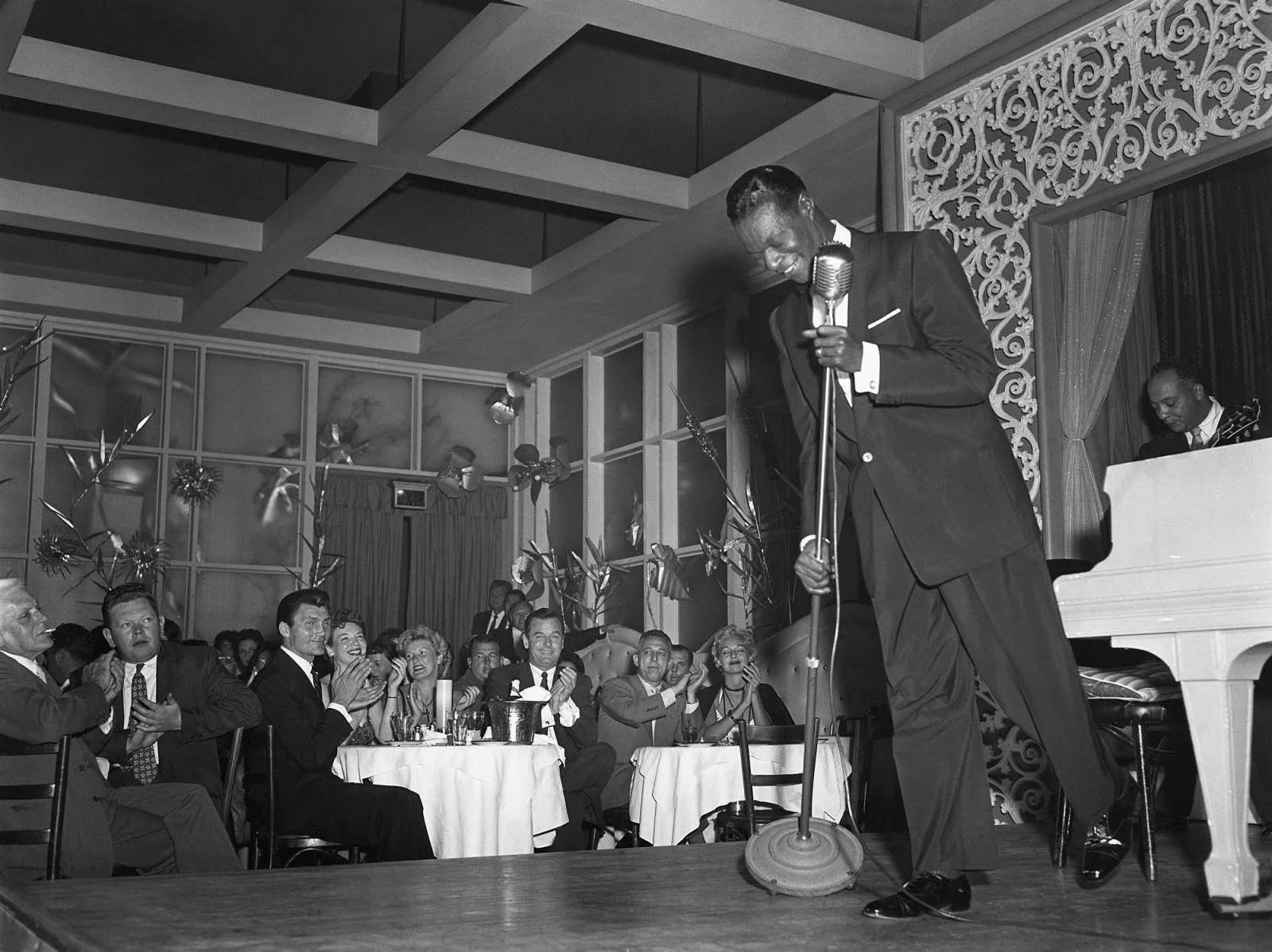 A black and white photo of a man in a tuxedo singing into a microphone on a stage, with a seated band member playing the piano and an audience clapping and enjoying the performance.