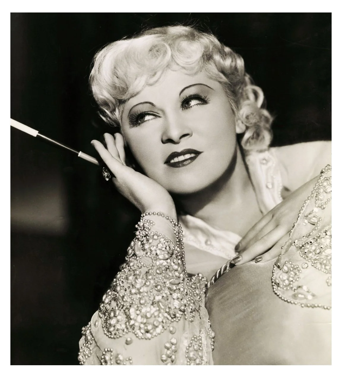 Black and white portrait of a glamorous woman with vintage hairstyle and makeup, wearing a beaded dress and jewelry, holding a cigarette holder near her face, with a thoughtful expression.