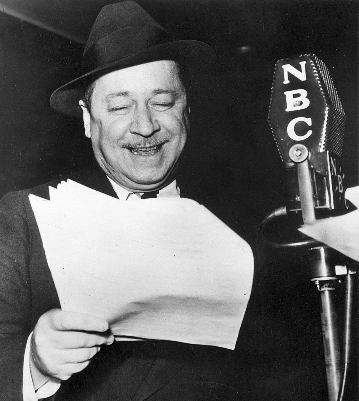 A black-and-white photo of a man wearing a suit and a tuxedo, reading from a sheet of paper into a vintage microphone with 'NBC' written on it, smiling and wearing a hat.