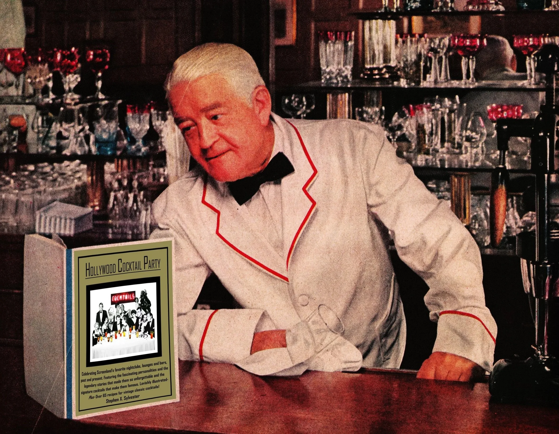 A man in a white tuxedo with red trim and a black bow tie, behind a bar with a variety of glassware in the background, leaning on the bar counter with a small sign that reads 'Hollywood Cocktail Party'.