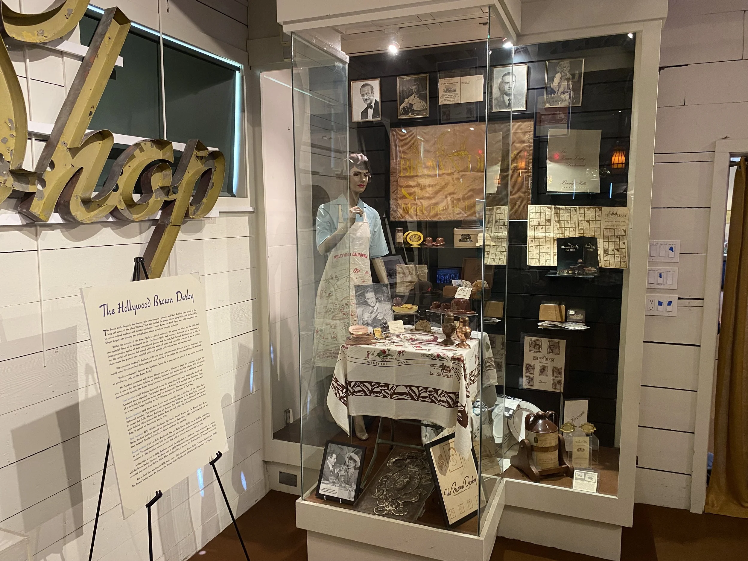 Display case with vintage items and a mannequin dressed in period clothing behind glass at a museum exhibit about The Hollywood Brown Derby.