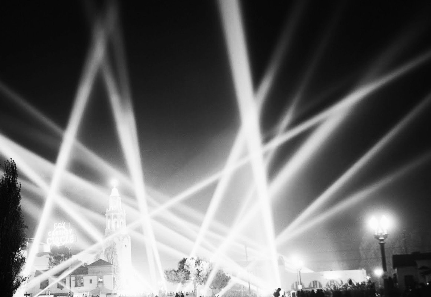 Night scene with multiple bright laser beams crossing in the sky, a clock tower in the background, trees, and a streetlamp on the right.