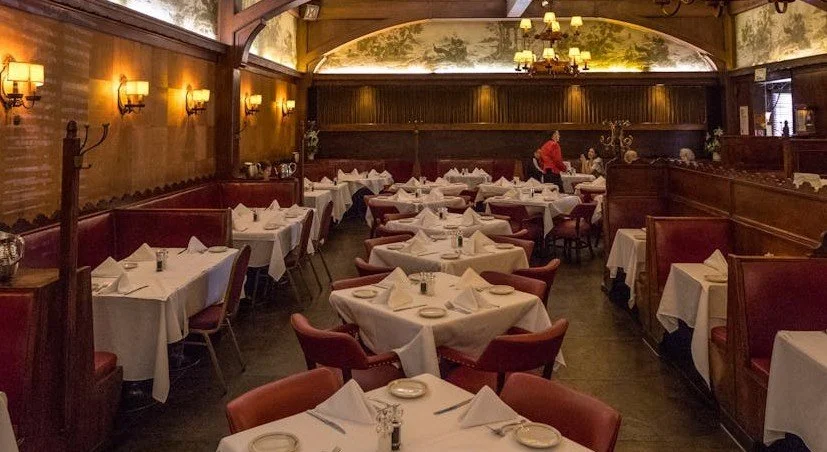 Elegant restaurant interior with tables set with white tablecloths, napkins, plates, and glassware, warm lighting, and wooden decor.