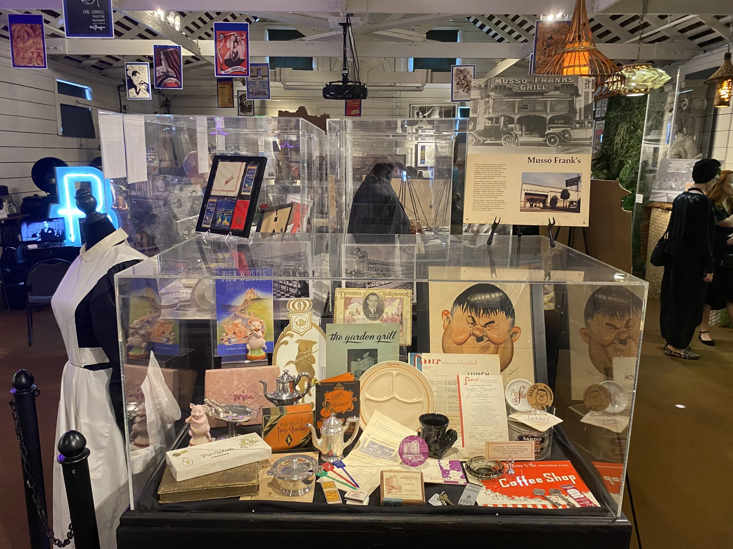 Display case containing vintage and nostalgic items such as posters, figurines, and memorabilia, with an informational sign titled "Musso Frank's" in the background, situated in a decorated room with hanging pictures and ambient lighting.