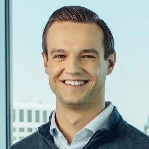 Smiling man in a professional setting with windows and cityscape in the background.