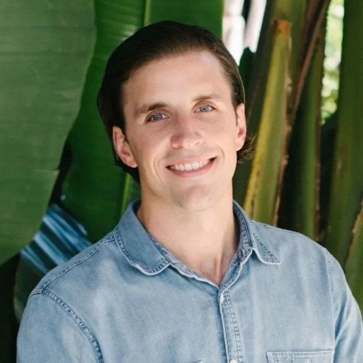 A young man with dark hair and light skin, smiling, wearing a light denim shirt, outdoors in front of large green tropical leaves.